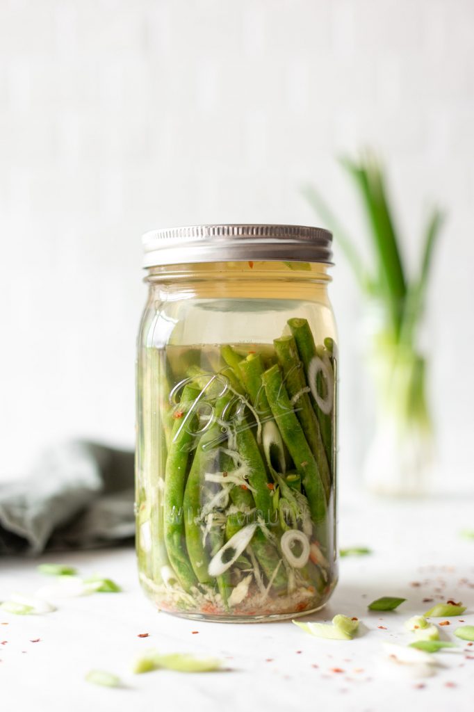 fermented green beans in a mason jar with scallion slices, grated ginger, and a salt brine. The green beans are held down in the brine with a glass fermentation weight