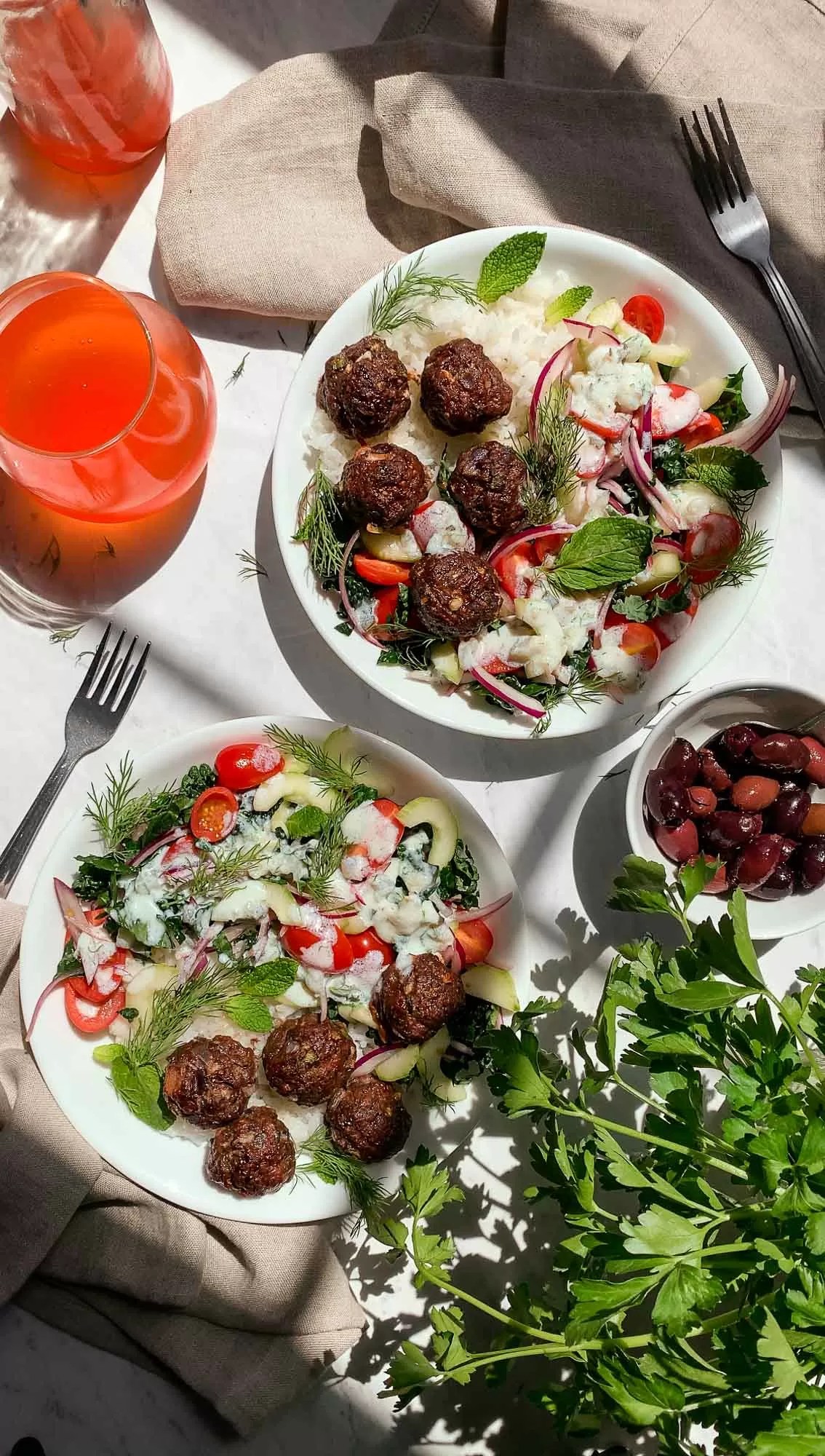 Two bowls on a table filled with rice, kale, tomatoes, cucumbers and red onion, topped with greek meatballs and drizzled with white Tzatziki sauce. 
