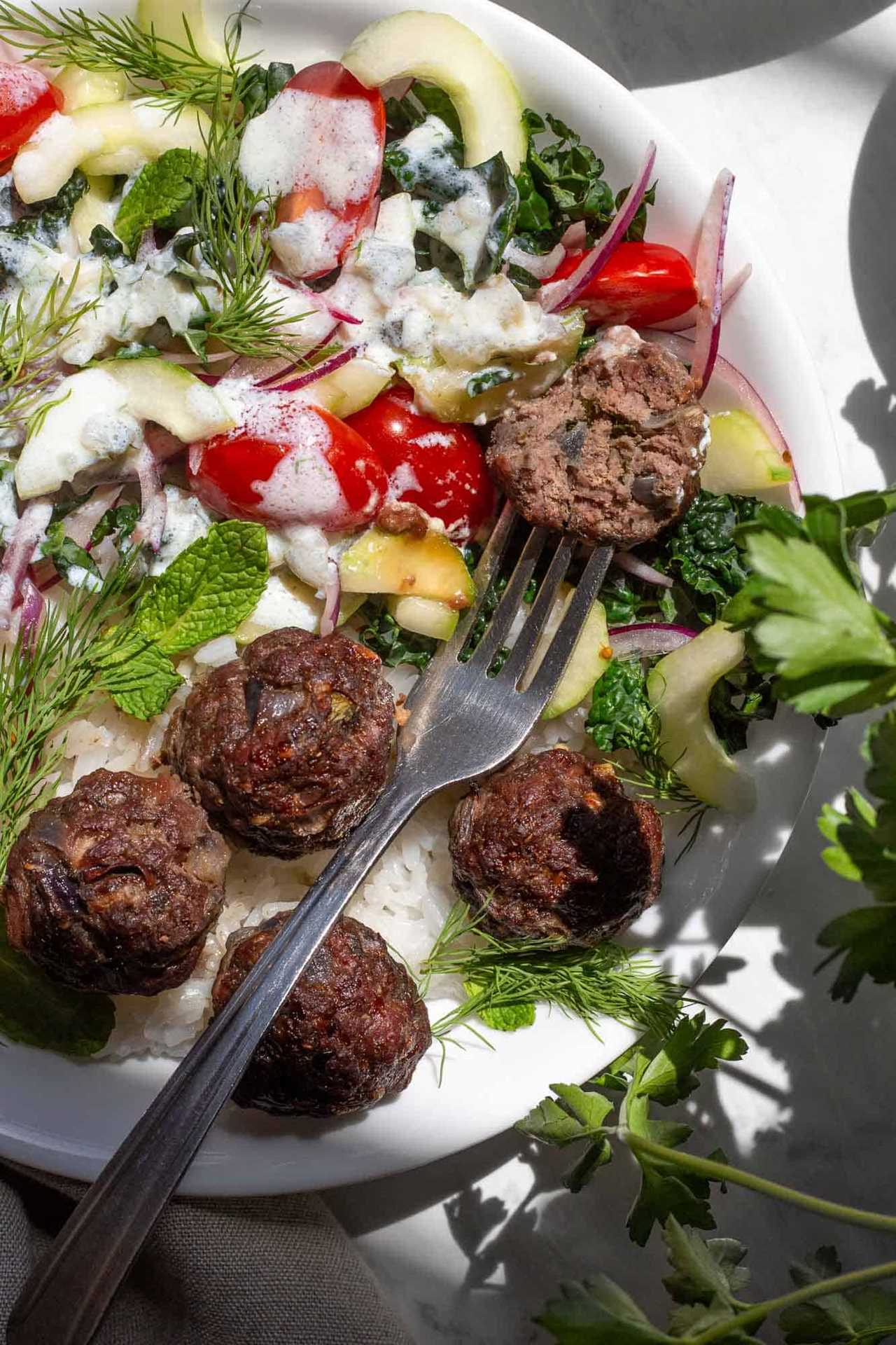a greek meatball with a bite taken out of it in a bowl with rice, vegetables and Tzatziki sauce.