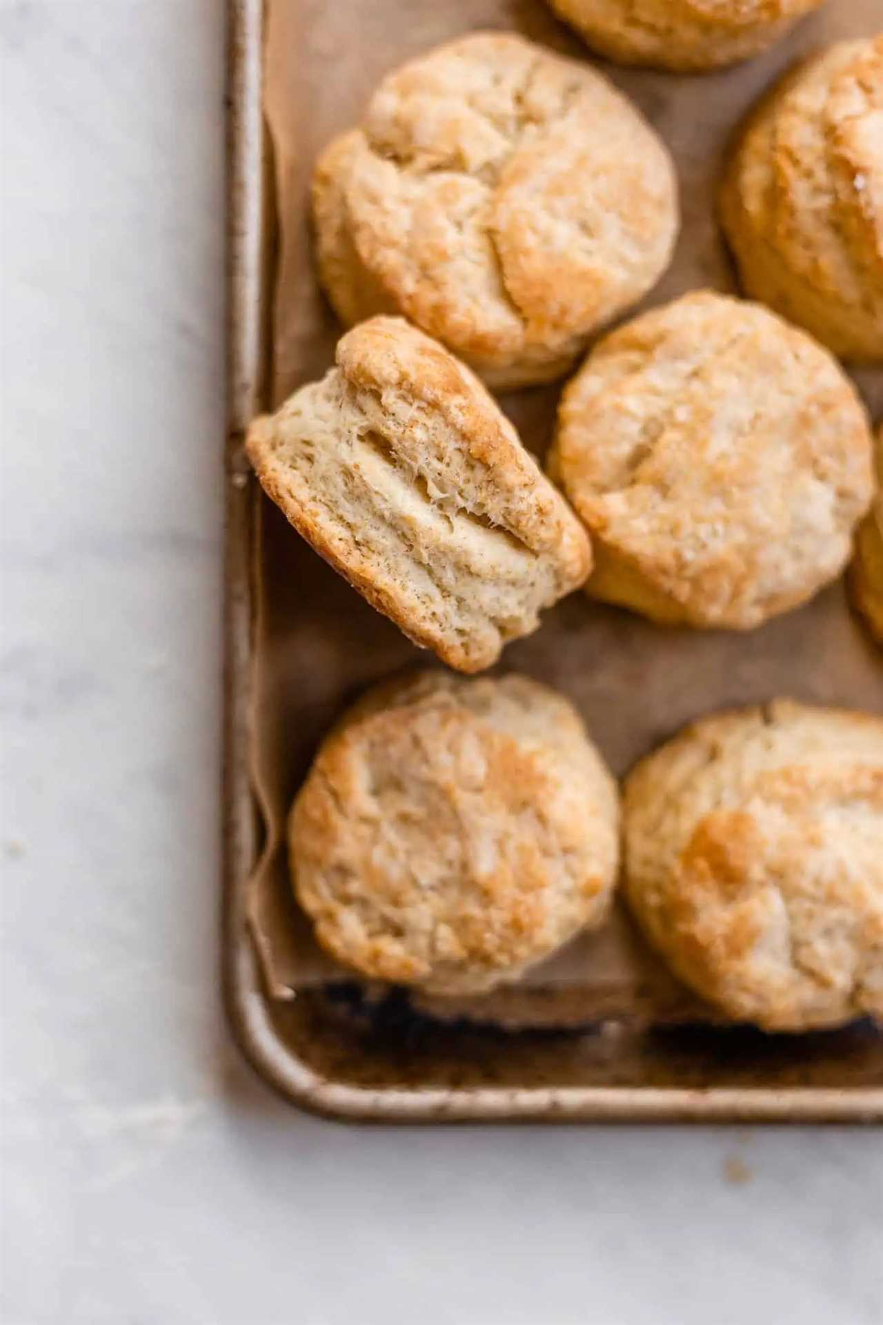 Golden, flaky sourdough biscuits on a sheet pan lined with parchment paper. Once biscuit is resting on its side to view the flaky, buttery layers. 