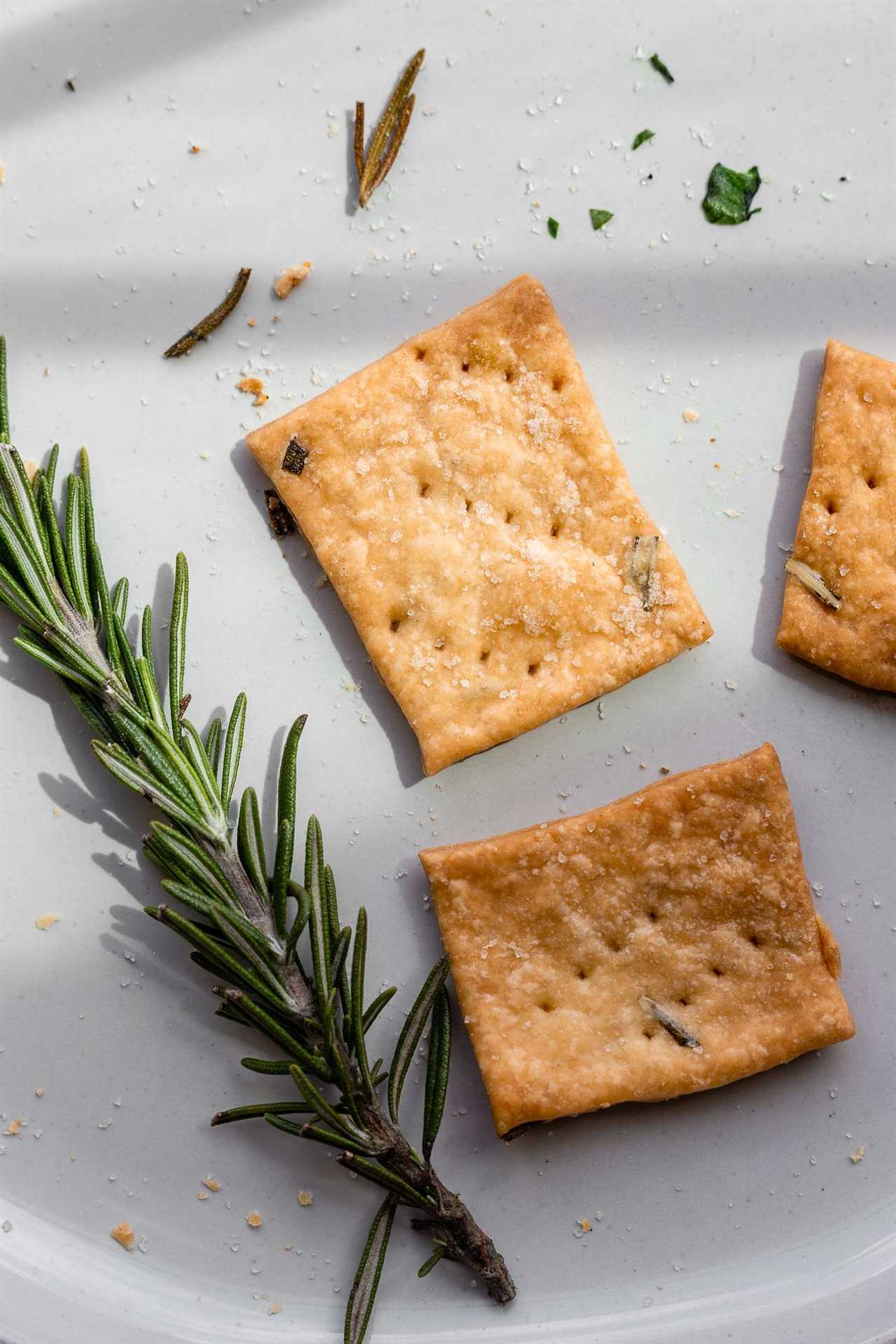 one crispy baked sourdough cracker on a plate next to fresh rosemary. Light hits the cracker to show textured sea salt and rosemary baked into the dough. 