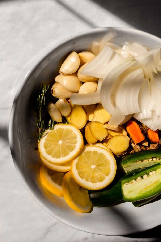 Fire Cider ingredients in a bowl, including chopped onion, garlic, ginger, turmeric, jalapenos, lemons and fresh sprigs of rosemary. 