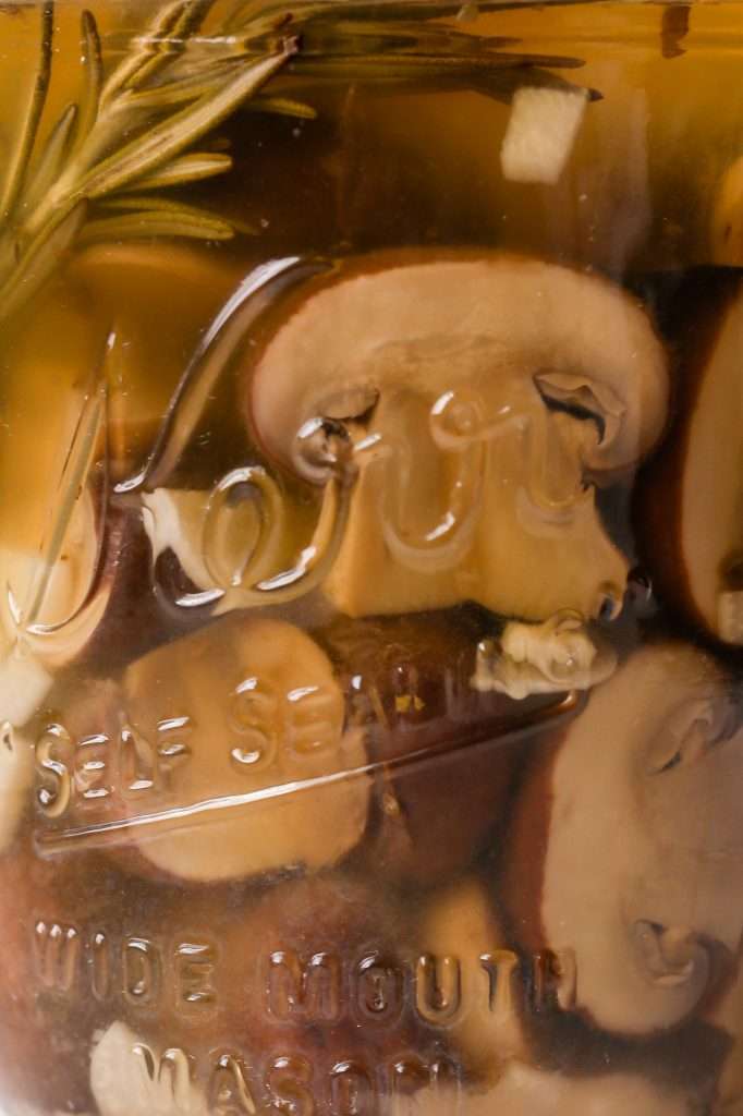 a close up of fermented mushrooms in a glass mason jar. The mushrooms and rosemary have fermented and changed to a muted brown color. 