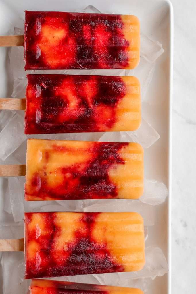 five cherry mango popsicles on a plate of ice. the popsicles are swirled with red cherry layers and yellow mango layers. 