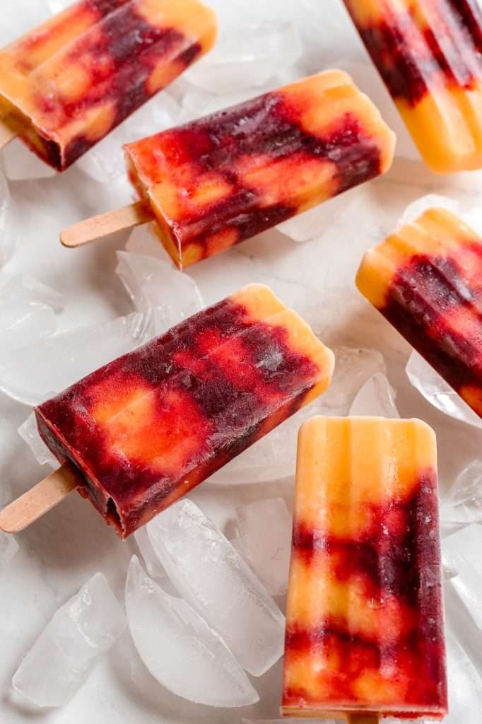 six cherry mango popsicles on a bed of ice. the popsicles are swirled with red cherry layers and yellow mango layers. 