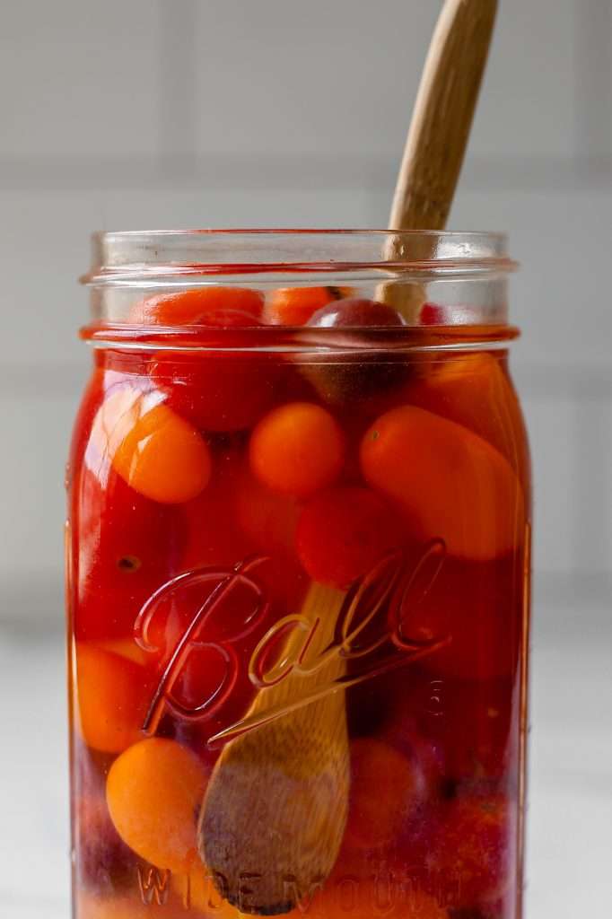 cherry tomatoes in a glass jar topped with sugar, water, and vinegar for fermenting. a wooden spoon rests in the jar, as the cherry tomato vinegar was just stirred. 