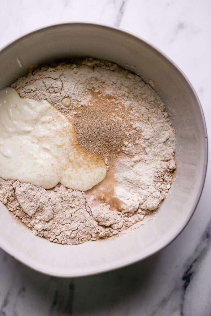 sourdough bread recipe without starter ingredients in a bowl. There's whole wheat flour, bread flour, instant yeast, yogurt, salt and water in a white bowl. 
