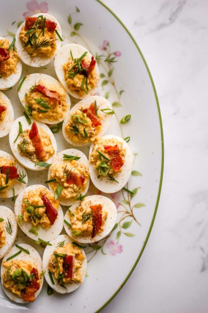 Jalapeno deviled eggs with bacon on a white plate that has a spring floral design on it. Each deviled egg is topped with a small crispy piece of bacon, fresh dill and minced green onion. 