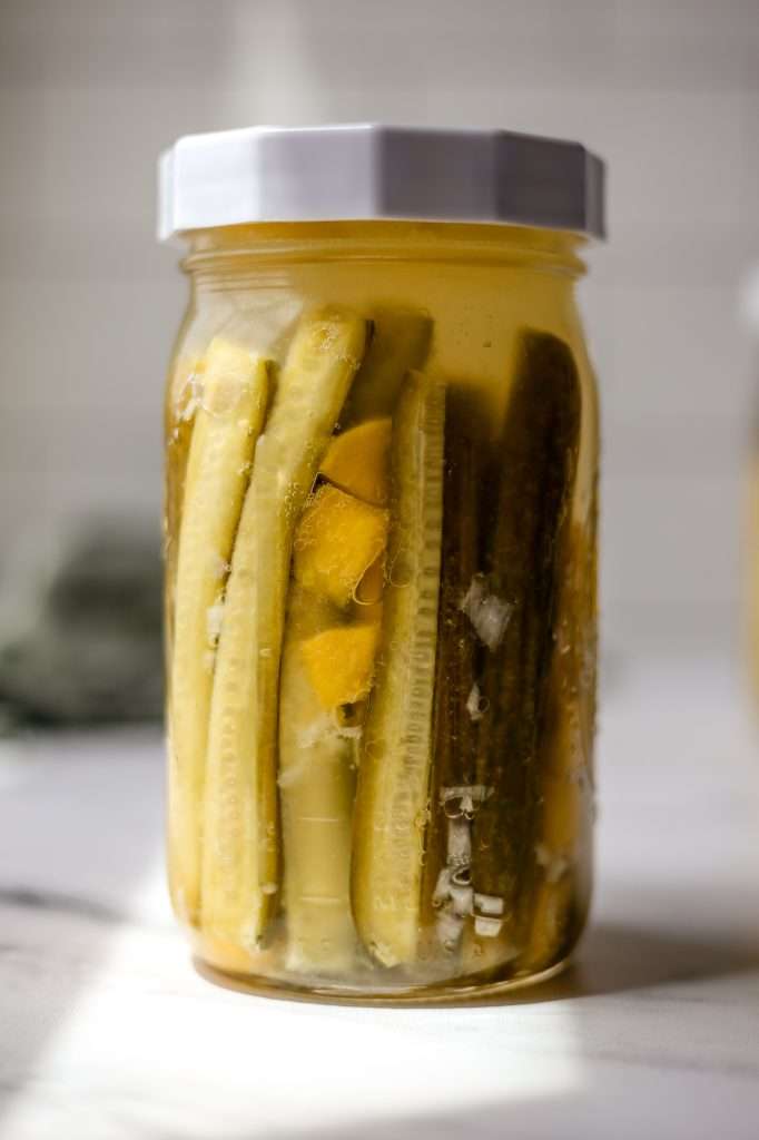 probiotic pickles in a large glass mason jar with a white lid. They are finished fermenting and the cucumbers have turned an olive green color. there are also fermented mango pieces in the jar with the pickles. 